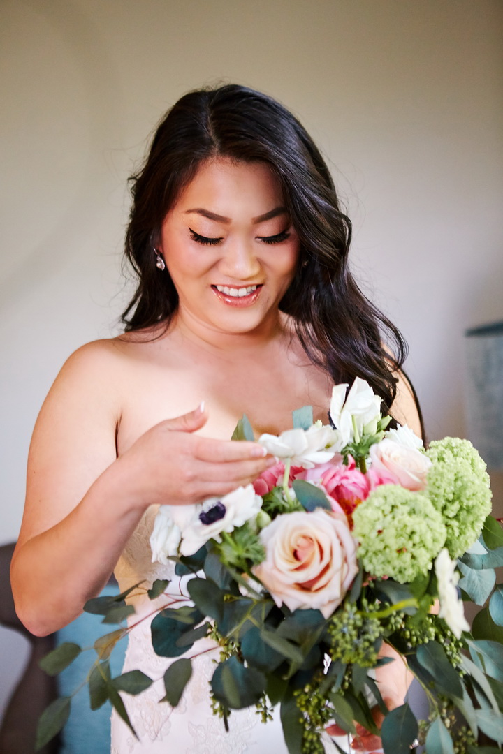 Bride smiling while looking down at bouquet of roses, anemones, and peonies.