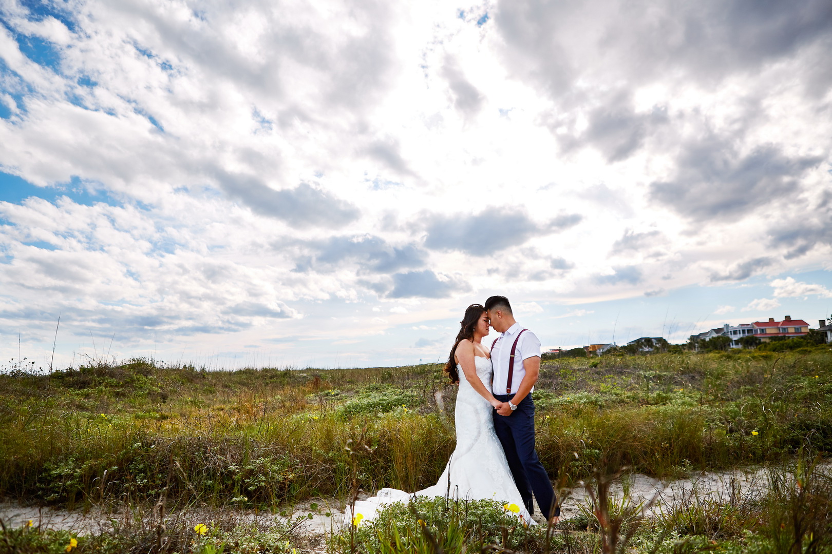 Bride and groom hold hands in beach grass, under dramatic clouds, on the beach in Isle of Palms, SC.