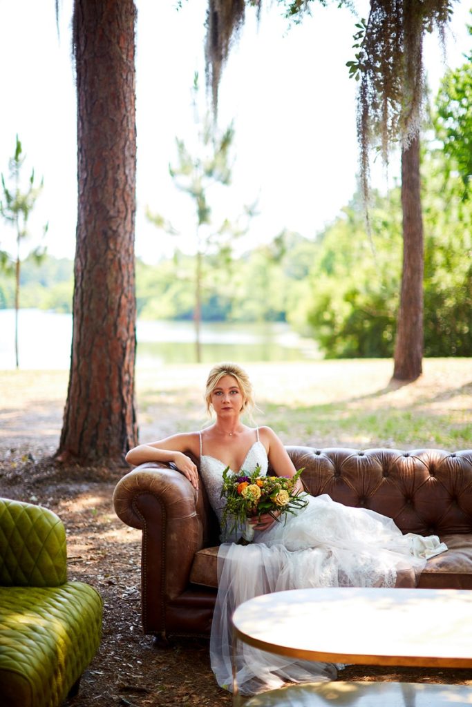 Bride lounging under oak tree, on brown leather couch, with colorful bouquet at Charleston Woodlands