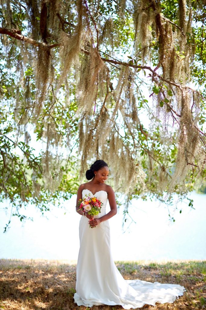 Bride looking over her shoulder with wedding dress, and colorful pink and orange bouquet, under oak trees and lakeside at Charleston Woodlands