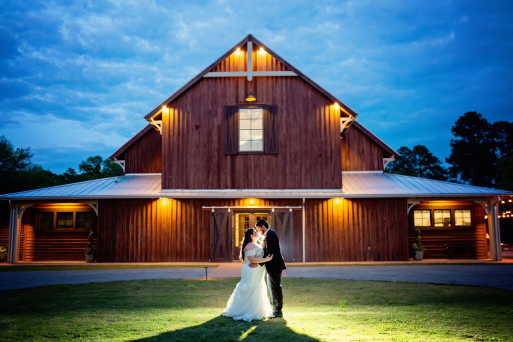 Bride and groom stand outside of the barn at Eden at Gracefield during dusk. They are backlit and embracing, and the barn is lit.  - photographed by Charleston wedding photographer Luxe By Lindsay Photography