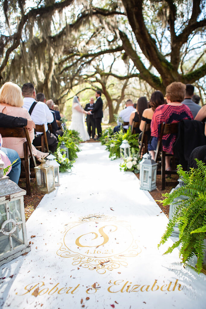 Personalized wedding aisle runner with monogrammed detail, with bride nd groom getting married in the background among oak trees at Eden at Gracefield - photographed by Charleston wedding photographer Luxe By Lindsay Photography