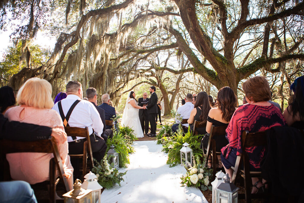 Wide photograph of wedding ceremony with bride and groom holding hands with oak trees in the background at Eden at Gracefield - photographed by Charleston wedding photographer Luxe By Lindsay Photography