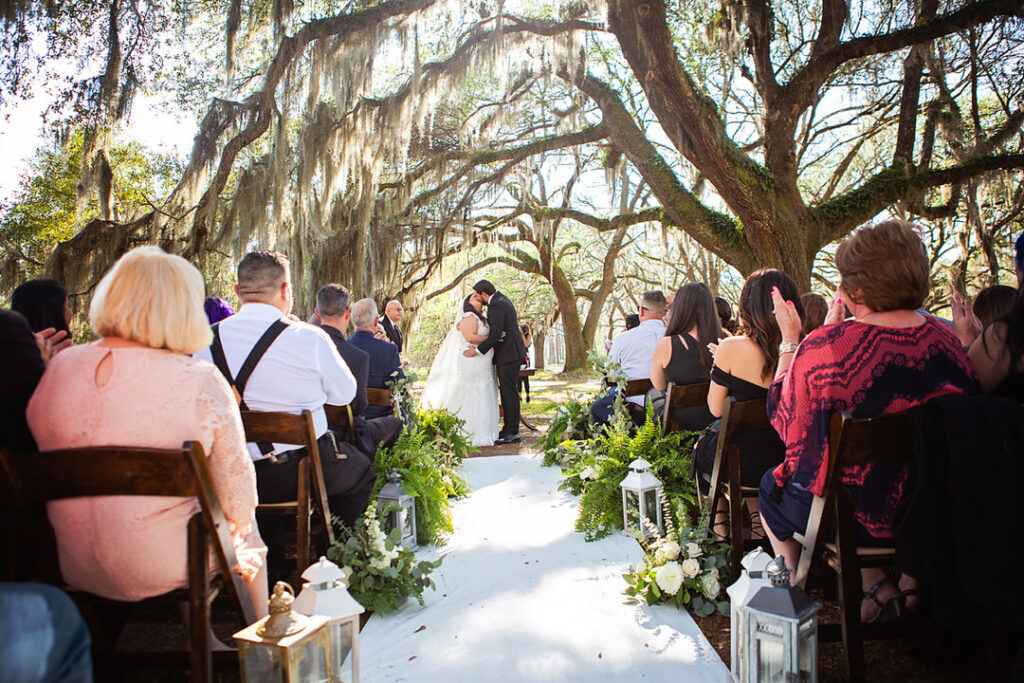 Bride and groom sharing their first kiss as a married couple, in a wide angle photo of their wedding ceremony with oak trees in the background  - photographed by Charleston wedding photographer Luxe By Lindsay Photography
