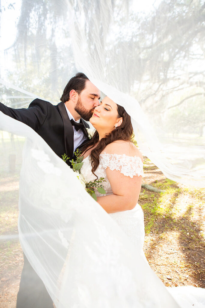 Groom kissing bride on the cheek while they are wrapped in the bride's windblown veil - photographed by Charleston wedding photographer Luxe By Lindsay Photography