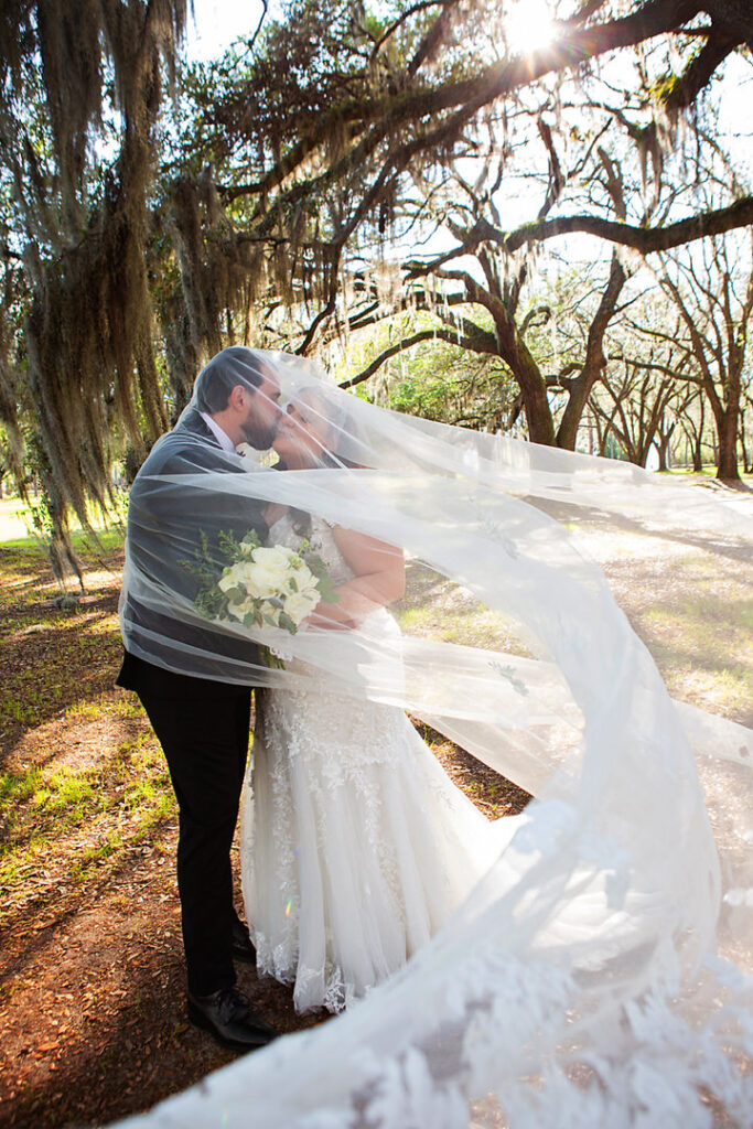 Full length portrait of bride and groom kissing underneath the bride's long wedding veil, with oak trees in the background at Eden at Gracefield - photographed by Charleston wedding photographer Luxe By Lindsay Photography