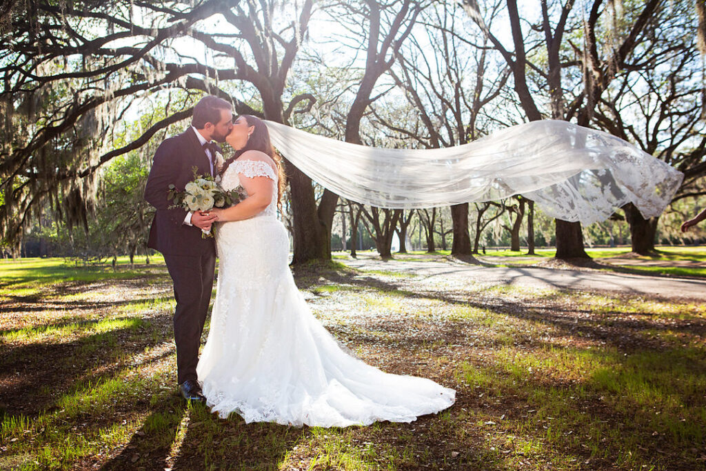 Bride and groom kiss while her veil is swept in the air by the wind, with oak trees in the background at Eden at Gracefield - photographed by Charleston wedding photographer Luxe By Lindsay Photography