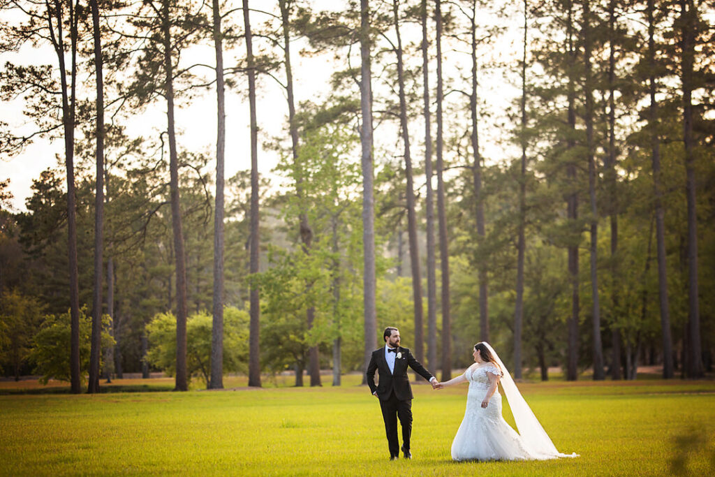 Wide angle photo of bride and groom looking at each other while holding. hands and walking on the lawn, with tall trees in the background at Eden at Gracefield - photographed by Charleston wedding photographer Luxe By Lindsay Photography