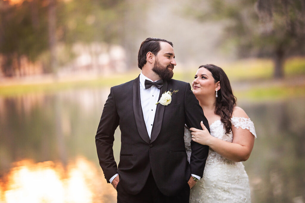 Bride holds onto groom's arm, while he has his hands in his pockets and they look at each other. Bride and groom are standing at the lake at Eden at Gracefield - photographed by Charleston wedding photographer Luxe By Lindsay Photography