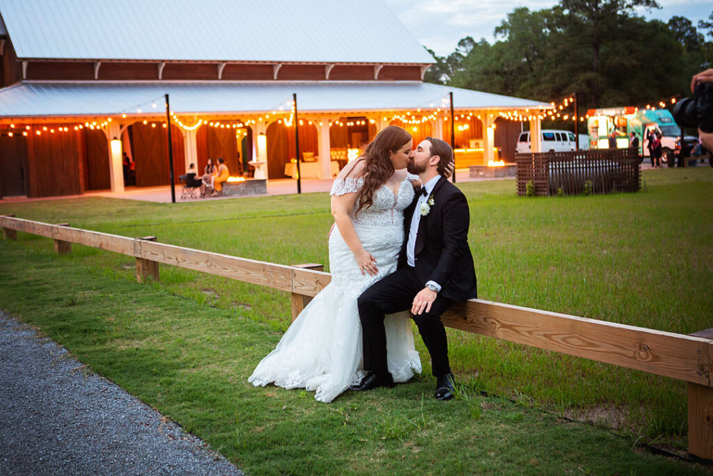 Bride and groom sit together and kiss with the lit reception barn in the background at Eden at Gracefield - photographed by Charleston wedding photographer Luxe By Lindsay Photography